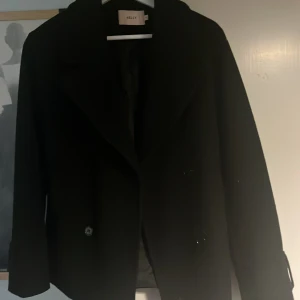 Svart rock från Nelly med knappar - Stilren svart rock från Nelly med klassisk krage och knäppning framtill. Jackan har en rak passform och långa ärmar, perfekt för dig som gillar en clean och tidlös look. Passar till många olika outfits och är enkel att matcha.