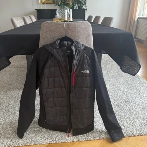 Svart pufferjacka från The North Face - Säljer en svart pufferjacka från The North Face med huva och quiltad framsida. Jackan har en röd dragkedja på bröstfickan och logga på både fram- och baksida. Tillverkad i polyester och syntetmaterial, perfekt för kyliga dagar.