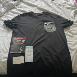 Svart Prada t-shirt med läderficka - Exklusiv svart t-shirt från Prada med en unik läderficka på bröstet, prydd med Pradas metallogo och en snygg dragkedja. T-shirten har korta ärmar och en klassisk rund hals. Materialet är mjuk bomull och detaljerna ger en lyxig vibe.