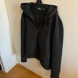 Svart cardigan jacka Polo Ralph Lauren - Svart hoodiejacka från Polo Ralph Lauren med quiltad front och huva. Jackan har dragkedja, långa ärmar i mjukt tyg och diskret logga på bröstet. Väldigt stilren.