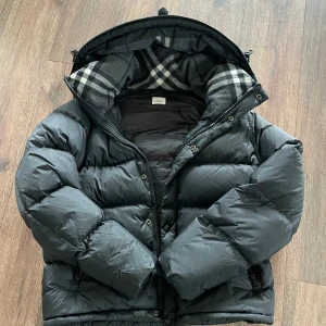 Svart dunjacka från Burberry - Säljer en svart dunjacka från Burberry med klassiskt rutigt foder i huvan och tydlig logga på bröstet. Jackan är tjock, har dragkedja och knappar framtill samt långa ärmar. Perfekt för kalla dagar och har en riktigt snygg streetstil.