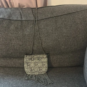 Silvrig axelväska med pärlor och fransar - Säljer en liten silvrig axelväska med pärlbroderi och dekorativa fransar längst ner. Väskan har ett unikt mönster och en tunn kedja som axelrem. Perfekt för att lyfta din outfit med lite extra bling.