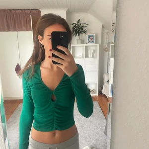 Grön croppad långärmad topp - Säljer en grön croppad topp med lång ärm och snygg v-ringning. Toppen har ett coolt rynkat parti framtill med en liten slits och håldetalj vid bröstet. Materialet är mjukt och stretchigt, perfekt för dig som gillar trendiga och bekväma plagg.