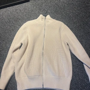 stickad kofta från Maison Margiela - Snygg beige stickad kofta från Maison Margiela med ribbad struktur och hel dragkedja framtill. Hög krage och långärmad modell som ger en clean och chill vibe. Perfekt för lager-på-lager och funkar till både jeans och dressade byxor.