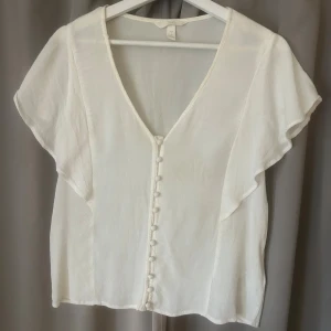 Vit blus med knappar och v-ringning - Säljer en söt vit blus från H&M med v-ringning och dekorativa tygklädda knappar framtill. Blusen har korta, luftiga ärmar. Använd fåtal gånger! 