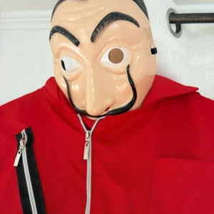 Röd La Casa de Papel-dräkt med mask - Säljer en röd jumpsuit inspirerad av La Casa de Papel, komplett med dragkedja framtill, stora fickor och tillhörande mask. Dräkten är i bomullsliknande material och har resår i midjan samt långa ärmar med elastiska muddar. Perfekt för temafest eller maskerad!