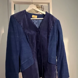 Blå kavaj från Anna Blomster design - Unik blå kavaj med snygga detaljer från Anna Blomster design. Jackan har en cool mix av material med mockaliknande partier och linnekänsla, samt dekorativa korsstygn längs axlarna. Stängs med knappar framtill och har lång ärm. 