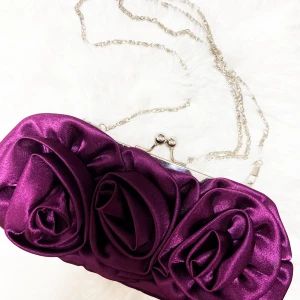 Lila kuvertväska med rosdetaljer - Kuvertväska i glansig lila satin med stora, tredimensionella rosdetaljer på framsidan. Väskan har en silverfärgad kedja och ett metallspänne. Insidan är fodrad med blommigt tyg i vitt, rosa och grönt. Perfekt för att lyfta din outfit med en unik touch.