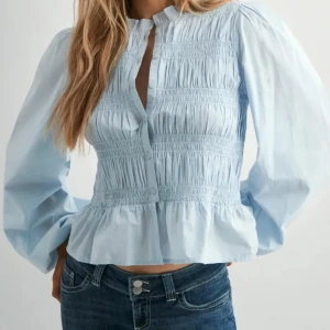 Ljusblå blus med volang och knappar - Säljer en ljusblå blus med smockad överdel, volang nedtill och knappar framtill. Blusen har långärmade puffärmar och en liten ståkrage. Perfekt för dig som gillar romantiska detaljer och en fräsch look.