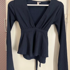 Svart omlottopp med knyt från Nelly - Säljer en marinblå omlottopp från Nelly i storlek XS. Toppen har ribbad struktur, lång ärm och snygg v-ringning. Knytband i midjan som kan knytas både fram och bak. Perfekt att styla med jeans eller kjol🙌🏼🙌🏼