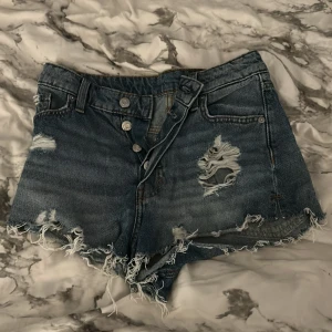 Blå high waist jeansshorts &Denim - Säljer ett par blå high waist jeansshorts från &Denim i modell Cheeky Shorts. De har slitna detaljer, fransig kant och flera snygga hål för en riktigt edgy look. Perfekta för sommaren och ger en cool vibe till din outfit.