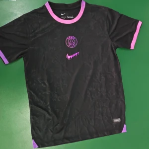 Svart PSG fotbollströja lila detaljer - Snygg svart PSG fotbollströja med lila detaljer på krage och ärmslut. Tröjan har korta ärmar och ett diskret blommönster i tyget. PSG-logga och Nike-märke i lila framtill. Tillverkad i lätt och ventilerande material. Ej officiell!!!
