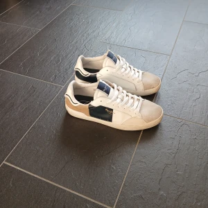 Pepe Jeans vita och blå sneakers - Snygga sneakers från Pepe Jeans i vitt med blå och beige detaljer. Skorna har rund tå, platt sula och klassisk snörning. Materialmix av skinn och mocka ger en cool kontrast. Perfekt för dig som gillar stilrena och sportiga sneakers med en twist. Storlek 42. In princip nya, bara använda ett fåtal gånger. Ska se lite slitna ut på sulan.