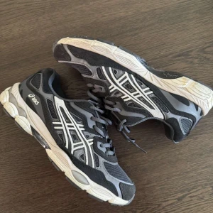 Svarta och grå Asics sneakers - Snygga Asics sneakers i svart och grått med vita detaljer och klassisk logga på sidan. Skorna har en rund tå, snörning och en tjock, vit sula med grå inslag. Materialet är en mix av mesh och syntet för bra komfort och andningsförmåga.