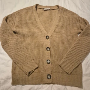 Brun stickad kofta från Pieces - Brun stickad kofta från Pieces med v-ringning och stora, snygga knappar framtill. Koftan har långärmad passform och är ribbstickad för en mysig och trendig look. Perfekt att slänga på sig när det är kyligt ute.