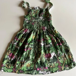 Grön djungelklänning från Kappahl Kids - Supersöt kort klänning med djungelmönster i grönt, rosa och gult från Kappahl Kids. Smock upptill och knytbara axelband. Mjukt bomullstyg och lekfulla detaljer med apor, krokodiler och växter. Perfekt för varma dagar och somriga äventyr.
