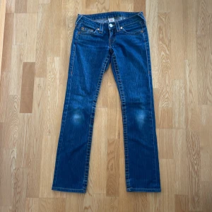 True Religion blå jeans, stl 26 - Säljer ett par klassiska blå jeans från True Religion med ikoniska bakfickor och kontrastsömmar. Storlek 26, normal benlängd 32. Modellen Johnny (utgått) har raka ben och mycket låg midja, tillverkade i slitstarkt denim. Snygga detaljer med logga på bakfickan. Liten slitning på höger ben vid knät, baktill (ej hål). 