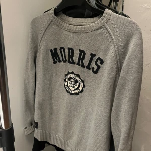 Grå stickad tröja från Morris - Snygg grå stickad tröja från Morris med svart broderad logga och emblem på bröstet. Tröjan har rund halsringning, ribbade muddar och långa ärmar. Perfekt för dig som gillar klassisk och stilren look.