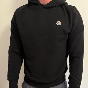 Svart hoodie från Moncler - Svart hoodie från Moncler med klassisk logga på bröstet. Tröjan har huva, långa ärmar och är tillverkad i mjuk bomull. Perfekt för dig som gillar stilrena och exklusiva plagg med en clean look. Storlek 3/ L och är helt ny aldrig använd endast testad! Nypris: 6000kr 