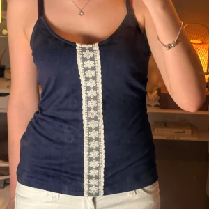 Mörkblå linnetopp med spets  - Supersöt mörkblå linnetopp från Love to Lounge med smala axelband som korsas i ryggen. Framtill har toppen en vit spetsdetalj med blommönster som ger en extra touch. Materialet är mjukt och stretchigt. 🥰