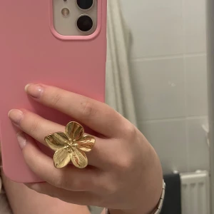 Stor guldfärgad blomring - Säljer en riktigt snygg och iögonfallande ring i guldfärg med en stor blomformad design. Perfekt för dig som vill sticka ut och addera lite extra bling till din outfit. Ringen har fem blad och en glansig finish som fångar ljuset.