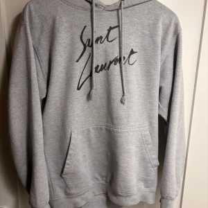 Grå Yves Saint Laurent hoodie med tryck - Snygg grå hoodie från Saint Laurent med svart logotyptryck framtill. Klassisk känguruficka, huva med snörning och långa ärmar. Perfekt för en chill och trendig look. Mjuk och skön i bomullsblandning.
