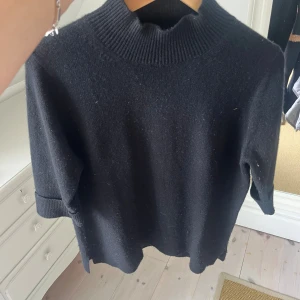 Svart polotröja i ull, kort ärm - Svart polotröja med ribbad hög krage och korta ärmar med uppvik. Tröjan är gjord i mjuk ull och har en loose passform som ger en chill vibe. Perfekt för lager-på-lager och funkar till både jeans och kjol. Snygg och enkel att styla.