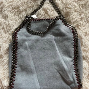 Grå axelväska från Stella McCartney - Snygg grå axelväska från Stella McCartney med grov silverkedja som axelrem och coola röda detaljer längs kanterna. Väskan har en mjuk yta och insidan är fodrad i rosa med logotyp. Perfekt för dig som gillar statement-accessoarer.