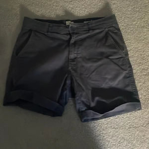 Två par shorts i bomull - Säljer två par stilrena shorts i bomull, ett par i mörkblått och ett par i beige. Båda har klassisk passform med fickor på sidorna, hällor för bälte och uppvikta benslut. Perfekta för varma dagar och enkel att matcha med t-shirt eller skjorta.