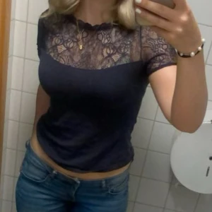 Svart spetsblus med kort ärm - Säljer en svart blus med korta ärmar och transparent spets upptill. Blusen har en rund halsringning med vågig kant och ett snyggt blommigt spetsmönster över axlar och bröst. Passformen är normal och materialet känns mjukt mot huden.