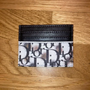Svart/Vit plånbok - Slimmad plånbok i svart och vitt med klassiskt logomönster. Plånboken har flera kortfack och är tillverkad i skinn med tryckt mönster. Perfekt för dig som vill ha en stilren och lyxig accessoar i fickan.