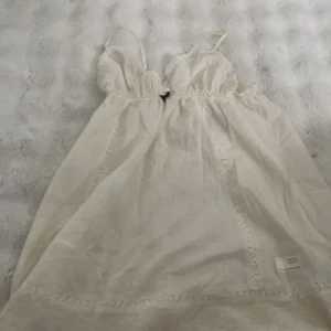 Odd Molly vit spetsklänning - Supersöt vit kortklänning från Odd Molly med spetsdetaljer, broderat motiv framtill och liten lila rosett vid bysten. Klänningen har tunna axelband, v-ringning och fina broderade kanter. Perfekt för sommarens alla tillfällen!