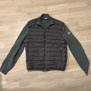 Grå Moncler dunjacka med stickade ärmar - Snygg grå dunjacka från Moncler med quiltad front och stickade ärmar och rygg. Jackan har dragkedja framtill, två sidofickor med dragkedja och Moncler-logga på ärmen. Perfekt för dig som vill ha en stilren och exklusiv look.