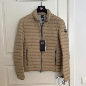 Beige dunjacka från Colmar (helt ny) - Säljer en snygg beige dunjacka från Colmar. Jackan har en quiltad design med dragkedja framtill och långa ärmar. Perfekt för kyligare dagar och ger en stilren look. Perfekt nu till våren. 