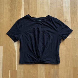 Snygg svart croppad t-shirt från H&M - Svart croppad t-shirt från H&M i 100% bomull. Toppen har korta ärmar, rund halsringning och en snygg knutdetalj framtill. Perfekt till jeans eller kjol för en chill och stilren look.
