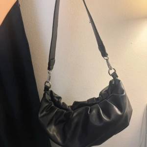 Svart axelväska i skinnimitation - Snygg svart axelväska med rynkad design och bred axelrem. Väskan har en mjuk, rundad form och är tillverkad i skinnimitation med silverfärgade metalldetaljer. Perfekt för dig som gillar trendiga accessoarer med lite edge.