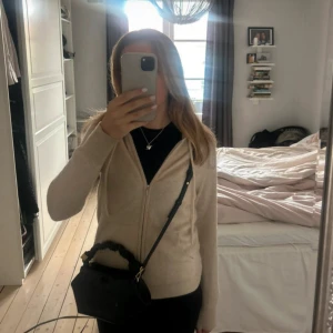 Beige hoodie i kashmir med dragkedja - Mysig beige hoodie i kashmir med dragkedja framtill och snörning i huvan. Tröjan har långa ärmar och två fickor framtill. Perfekt för dig som gillar en enkel och stilren look med lyxig känsla.