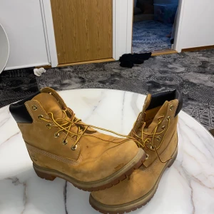 Timberland boots i mocka, gulbruna - Klassiska Timberland boots i gulbrun mocka med robust snörning och svart vadderad kant runt ankeln. Grov sula för extra grepp och ikonisk logga på sidan. Perfekta för dig som gillar streetstyle och vill ha en tidlös look.