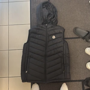 Svart dunväst från Moncler med huva - Svart dunväst från Moncler med snyggt quiltad design och huva. Västen har dragkedja framtill, två fickor med dragkedja och Moncler-logga på bröstet. Perfekt för lager-på-lager och streetstyle. Materialet är lätt och värmande, med en clean look.