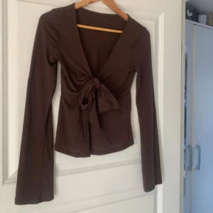Brun omlottopp med knyt från Gina Tricot - Supersnygg brun omlottopp från Gina Tricot med lång ärm och knyt framtill. Toppen har en djup v-ringning och är figurnära med lite croppad passform. Tillverkad i mjuk bomull som känns skön mot huden. Perfekt för dig som gillar trendiga och stilrena plagg. Använd 2 gånger så den är i väldigt fint skick😌❤️