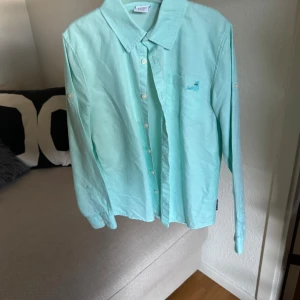 Mintgrön skjorta från Polarn O. Pyret - Fräsch mintgrön skjorta från Polarn O. Pyret med klassisk krage och vita knappar. Skjortan har lång ärm och liten broderad logga på bröstet. Perfekt för dig som gillar stilrena och ljusa plagg. Tillverkad i mjuk bomull.