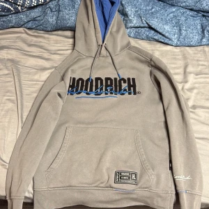 Grå hoodie från Hoodrich XS - Säljer en grå hoodie från Hoodrich i storlek XS. Tröjan har blått foder i huvan, broderad logga i svart och blått framtill, samt känguruficka och coola streetwear-detaljer. Perfekt för dig som gillar urban stil och snygga loggor.