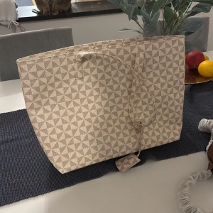 Beige shoppingväska med geometriskt mönster - Stor och rymlig shoppingväska i beige med stilrent geometriskt mönster. Väskan har snörning upptill och är tillverkad i ett kraftigt tygmaterial. Perfekt för dig som vill ha plats för allt du behöver under dagen. Finns ett mindre fack inuti. Aldrig använd 