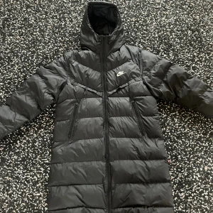 Svart lång dunjacka från Nike - Säljer en svart lång dunjacka från Nike med huva och dragkedja framtill. Jackan har två snedställda fickor med dragkedja och diskret Nike-logga på bröstet. Perfekt för kalla dagar med sin vadderade design och stilrena look. Har dock ett litet hål på ena sidan av arméns , men inget synligt!! Pris går att diskuteras vid snabb affär.