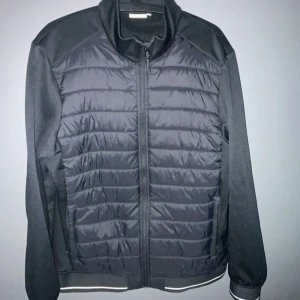 S.C.W cardigan - Svart cardigan från S.C.W. i storlek L med quiltad front och släta ärmar. Jackan har dragkedja framtill, ståkrage och diskreta reflexdetaljer vid muddar. Perfekt för dig som gillar en stilren och sportig look.