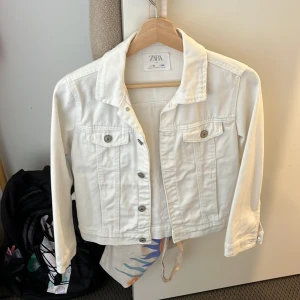 Vit jeansjacka från Zara - Snygg vit jeansjacka från Zara med klassisk krage, knappar framtill och två bröstfickor med lock. Jackan har lång ärm och är i ett stilrent, croppat snitt som passar perfekt till vår och sommar. Enkel att matcha med det mesta för en fräsch look. Använd 1 gång