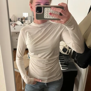 Beige långärmad topp med drapering hm - Säljer en beige långärmad topp med draperad effekt och hög hals. Toppen är tight och har en mjuk, stretchig känsla som sitter snyggt mot kroppen. Perfekt att styla med jeans eller kjol för en clean look.