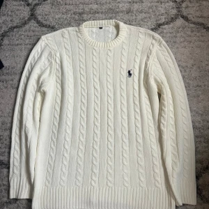 Vit kabelstickad tröja från Ralph Lauren - Kabelstickad vit tröja från Ralph Lauren med klassisk rund halsringning och broderad marinblå logga på bröstet. Tröjan har långa ärmar och ribbade muddar vid ärmslut och nederkant. Perfekt för en stilren och tidlös look.