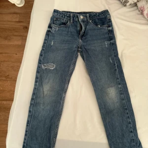 Blå jeans med slitningar från H&M - Säljer ett par blå jeans från H&M i relaxed fit med justerbar midja och avsmalnande ben. Jeansen har coola slitningar och en rippad detalj på ena benet. Perfekta för en avslappnad och trendig look.