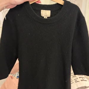 Svart kashmirtröja från H&M - Mjuk och lyxig svart tröja i kashmir från H&M. Tröjan har rund halsringning, ribbade muddar och kortare ärmar som slutar strax ovanför armbågen. Perfekt att styla med jeans eller kjol för en clean look.